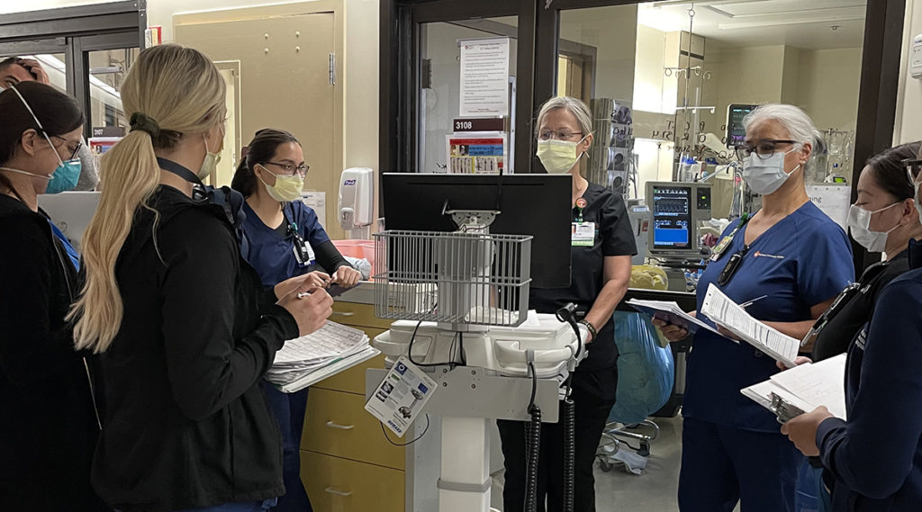 Multiple nurses in scrubs are huddled around a computer and are talking to each other