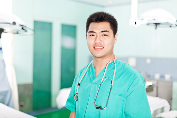 A nurse wearing bright green scrubs with a stethoscope around his neck. He stands in front of an operating room.