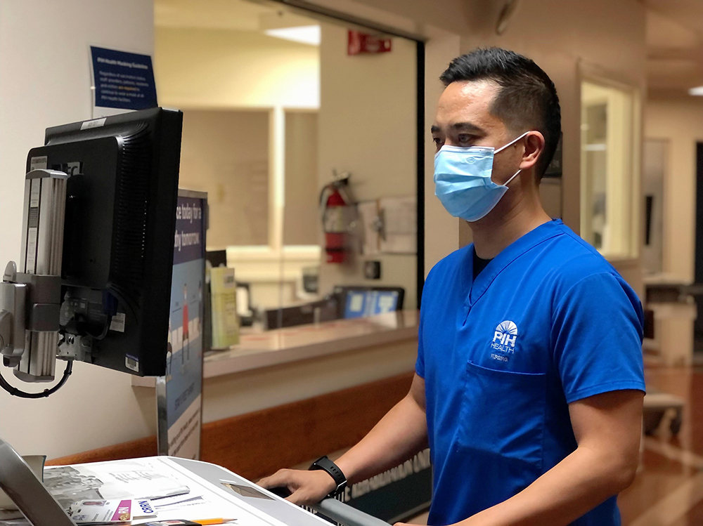 Shannon Bocalbos wearing scrubs and a mask stands in front of a computer he is typing on