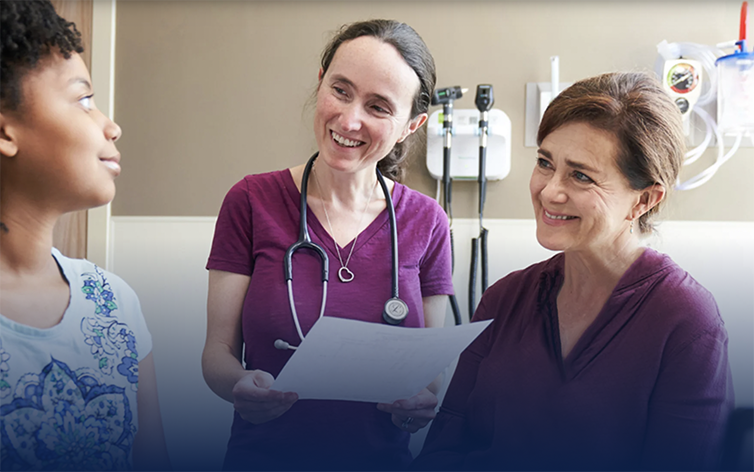 Three nurses are talking. Two are in scrubs and one is in casual attire.