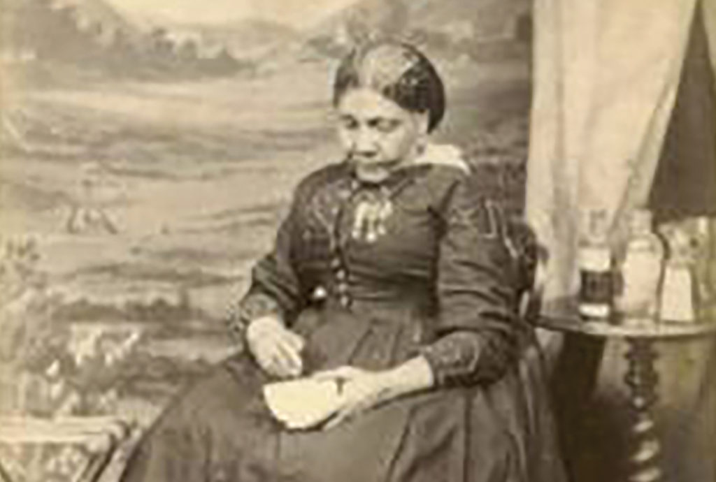 A sepia toned photo of Mary Seacole wearing a dress and sitting on a chair, looking down