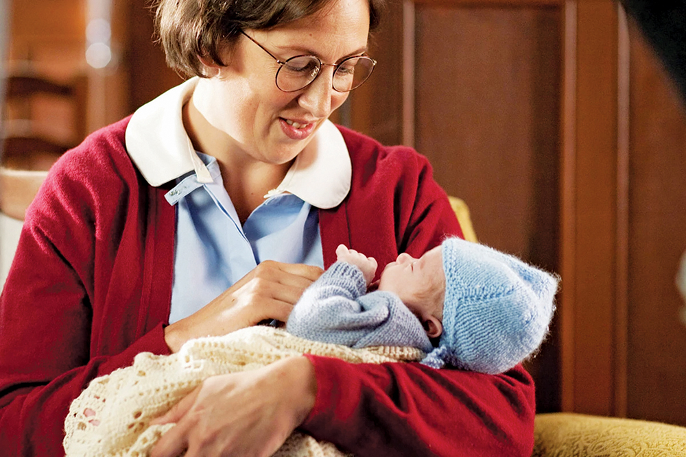 Chummy Browne from "Call the Midwife" is sitting and looking at a baby that she is holding in her arms.