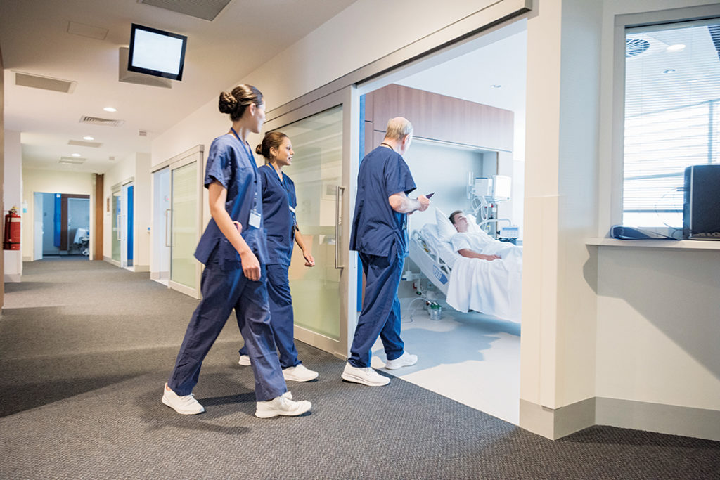 Three registered nurses in blue scrubs are walking into a patient's room