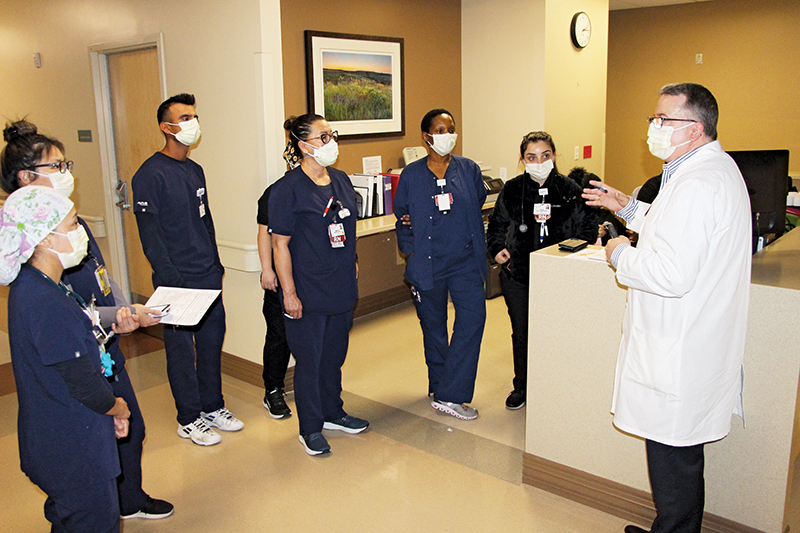 Male nurse wearing a mask and white coat addresses his team of nurses circled around him.
