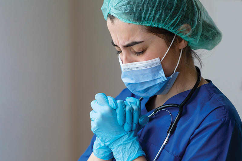 Nurse clasping hands and looking stressed while wearing a surgical cap, mask, and latex gloves.