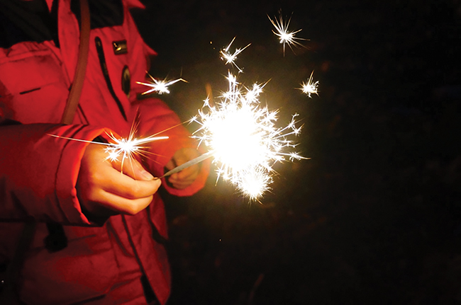 A child in a red coat holds a lit fireworks sparkler with his fingertips.