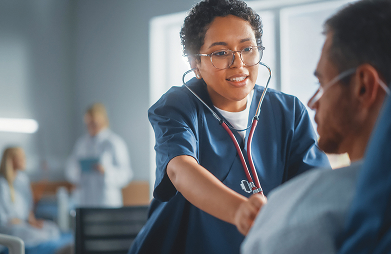 Nurse in blue scrubs listens to patient's heart with a stethoscope.