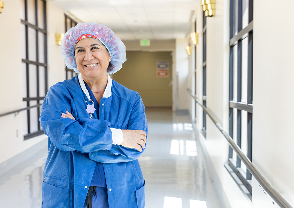 OR nurse Katie Curren smiling with arms crossed, wearing blue scrubs and plastic surgical cap standing in white hospital hallway.