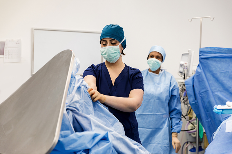 Operating room nurse wearing blue scrubs, a mask, and cap, discards her surgical gown into the trash bin.