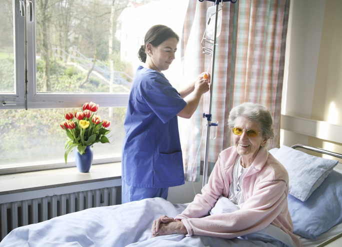 Nurse in hospital room opening the curtains while elderly patient in bed smiles.