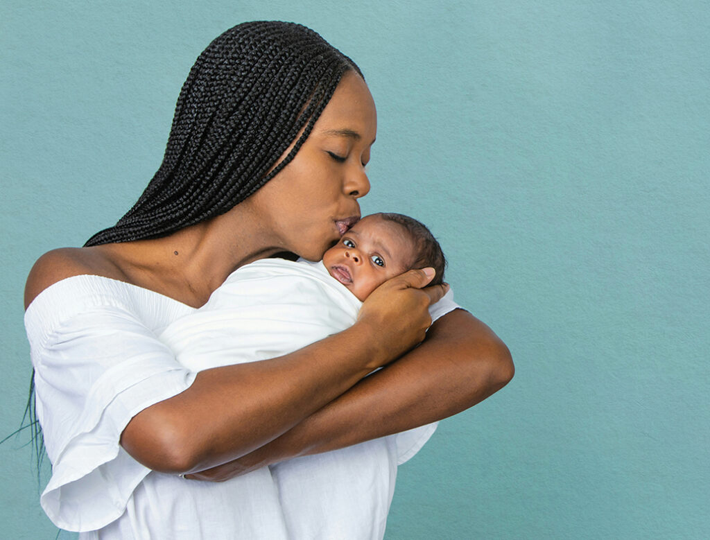 A beautiful young African-American woman with braids is kissing her newborn son and looking at him with love on a teal blue background.