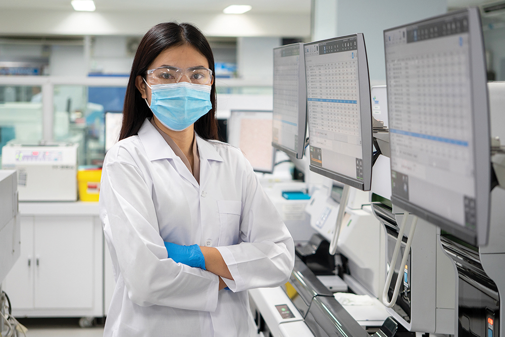 Nurse researcher in white coat and blue masks stands in a science lab with arms crossed.