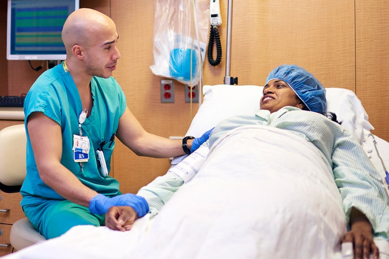 Male nurse sits by a patient's bed holding her hand.