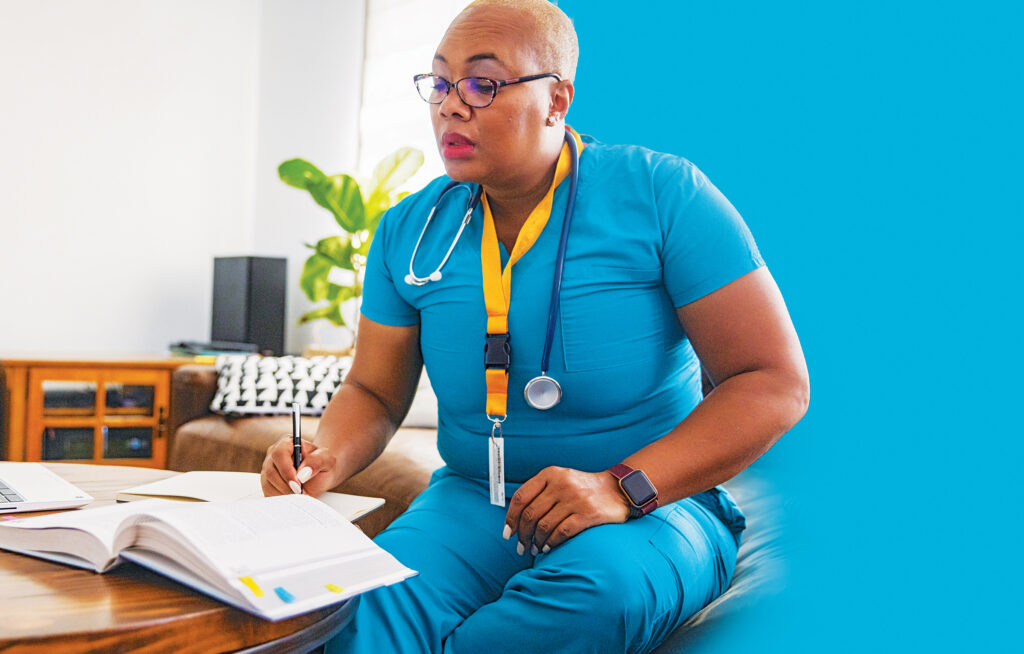 Nurse in blue scrubs sitting on a desk reading a textbook.