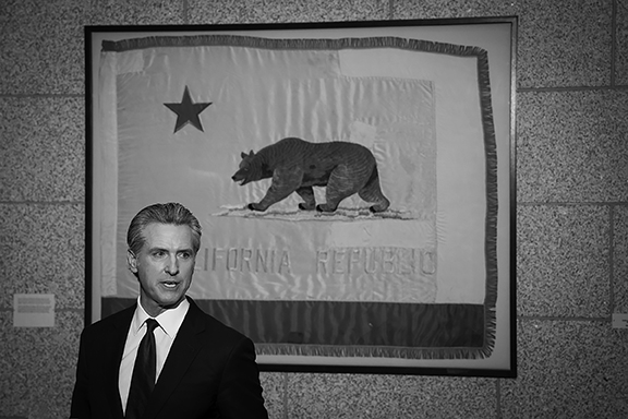 Black and white of Calif Gov. Gavin Newsom in front of state flag